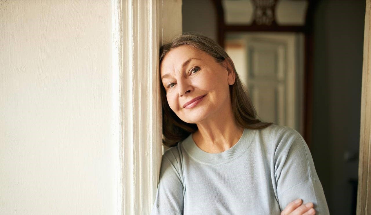 Attractive middle aged Caucasian woman with loose gray hair and wrinkles spending day indoors, leaning on white wall and smiling at camera.